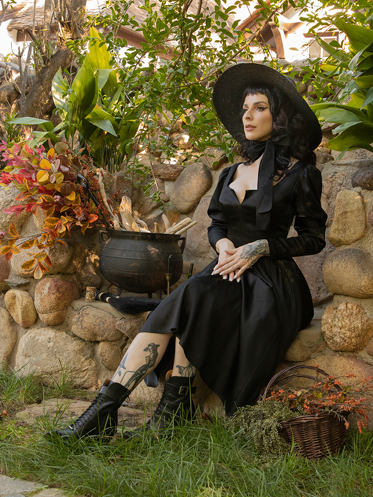 Getting ready to cast a spell, Stephanie sits next to a cauldron and wicker basket while wearing the latest goth dress from La Femme en Noir.