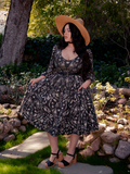 Immersed in the enchanting forest setting, a stunning brunette muse enthralls all with her presence, clad in the exquisite Dark Forest Dress embellished with the captivating Cottage Witch Toile Print, a gothic masterpiece hailing from La Femme en Noir.