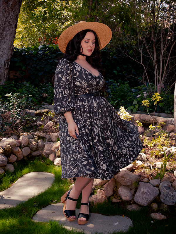 Amidst the whispers of the ancient woodland, a bewitching brunette figure entices with her grace, enveloped in the captivating Dark Forest Dress adorned in the enchanting Cottage Witch Toile Print, an exquisite creation by La Femme en Noir.