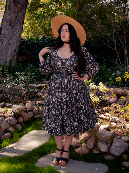 Amidst the haunting allure of the forest, an enchanting brunette model enchants with her poise, donning the mesmerizing Dark Forest Dress in the Cottage Witch Toile Print, a haunting creation by La Femme en Noir.