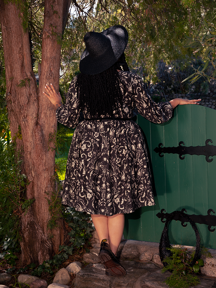 In the heart of the mystical woodland, an enchanting brunette figure enchants with her ethereal presence, donning the captivating Dark Forest Dress adorned with the mesmerizing Cottage Witch Toile Print, an alluring creation by La Femme en Noir, the goth dress artisan.