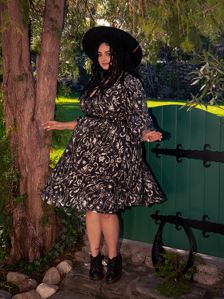 Amongst the ghostly foliage of the forest, a striking brunette model casts a spell with her presence, wearing the alluring Dark Forest Dress featuring the hauntingly beautiful Cottage Witch Toile Print, a creation by La Femme en Noir, the maestro of gothic fashion.