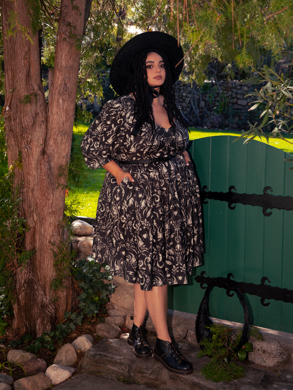 Enveloped in the haunting forest's embrace, a stunning brunette muse emerges, emanating an otherworldly aura in the Dark Forest Dress adorned with the ethereal Cottage Witch Toile Print, a mesmerizing ensemble crafted by goth dress maker La Femme en Noir.