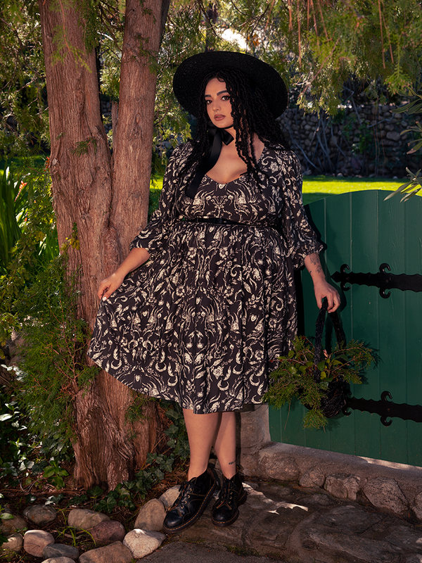 Within the eerie depths of the forest, a breathtaking dark-haired beauty captivates the senses, draped in the spellbinding Dark Forest Dress embellished with the enchanting Cottage Witch Toile Print, a masterpiece originating from the gothic couturier La Femme en Noir.