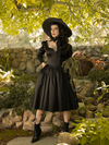 Holding a wicker basket full of picked flowers, Stephanie models the Cottage Witch Dress in Japanese Black Satin from gothic clothing make La Femme en Noir.