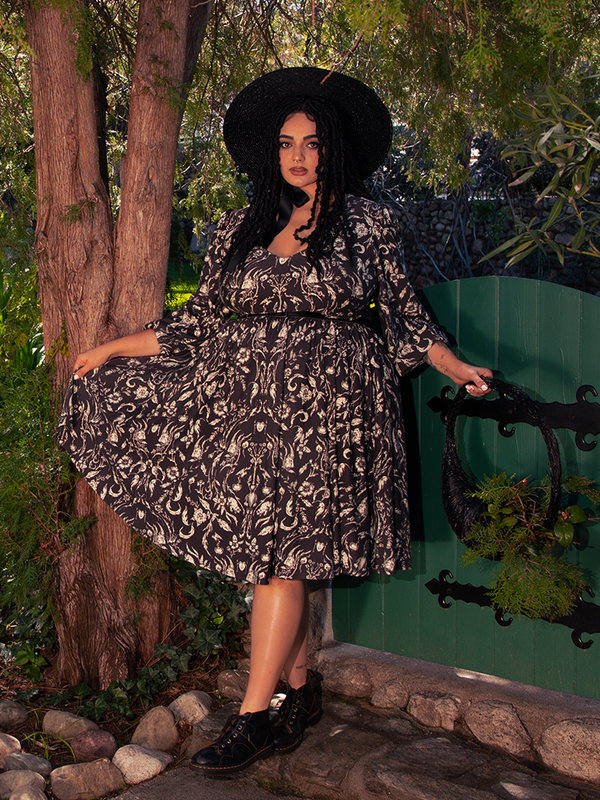 In the depths of the enigmatic woods, a mesmerizing brunette figure appears, captivating all with her aura, garbed in the beguiling Dark Forest Dress adorned with the captivating Cottage Witch Toile Print, a bewitching creation by La Femme en Noir.