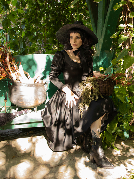 Stephanie Joens sitting on a bench next to a small cauldron of dead branches looking very gothic retro in her Cottage Witch Dress in Japanese Black Satin.