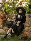 Sitting next to a cauldron filled with dead branches, Stephanie wears the latest goth dress release from La Femme en Noir - the Cottage Witch Dress in Japanese Black Satin.