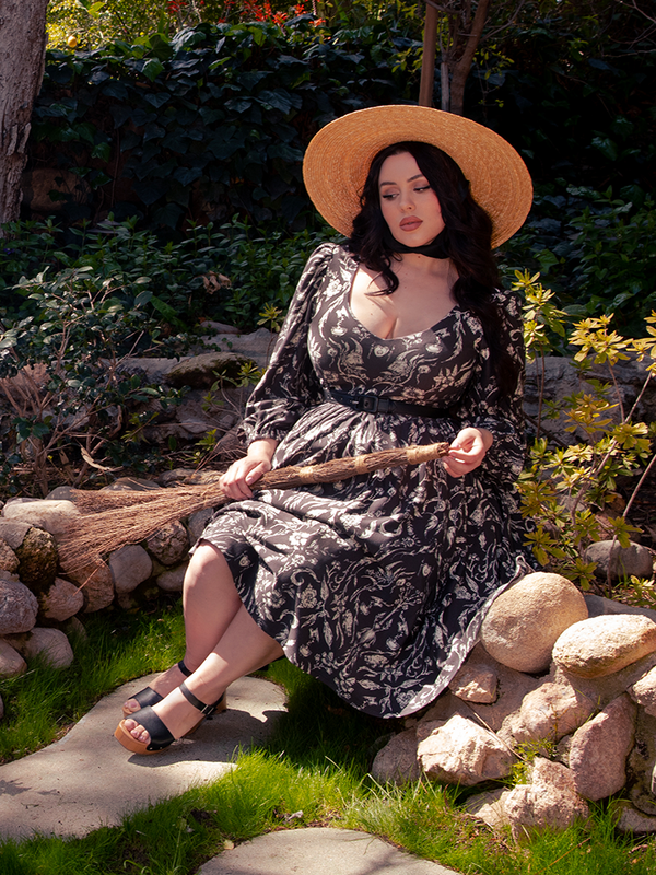 Lost in the enigmatic forest, a captivating brunette figure emerges, donning the spellbinding Dark Forest Dress adorned in the mystical Cottage Witch Toile Print, an enchanting design by goth dress maker La Femme en Noir.