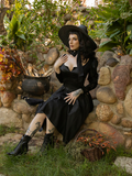 Stephanie Anne Joens sits on a rocky area adorned with plants while wearing the Cottage Witch Dress in Japanese Black Satin from La Femme en Noir.