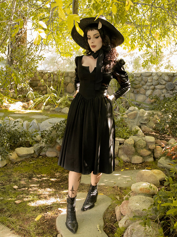 Walking on a stone pathway amongst a garden setting, Stephanie models the Cottage Witch Dress in Japanese Black Satin from goth dress maker La Femme en Noir.