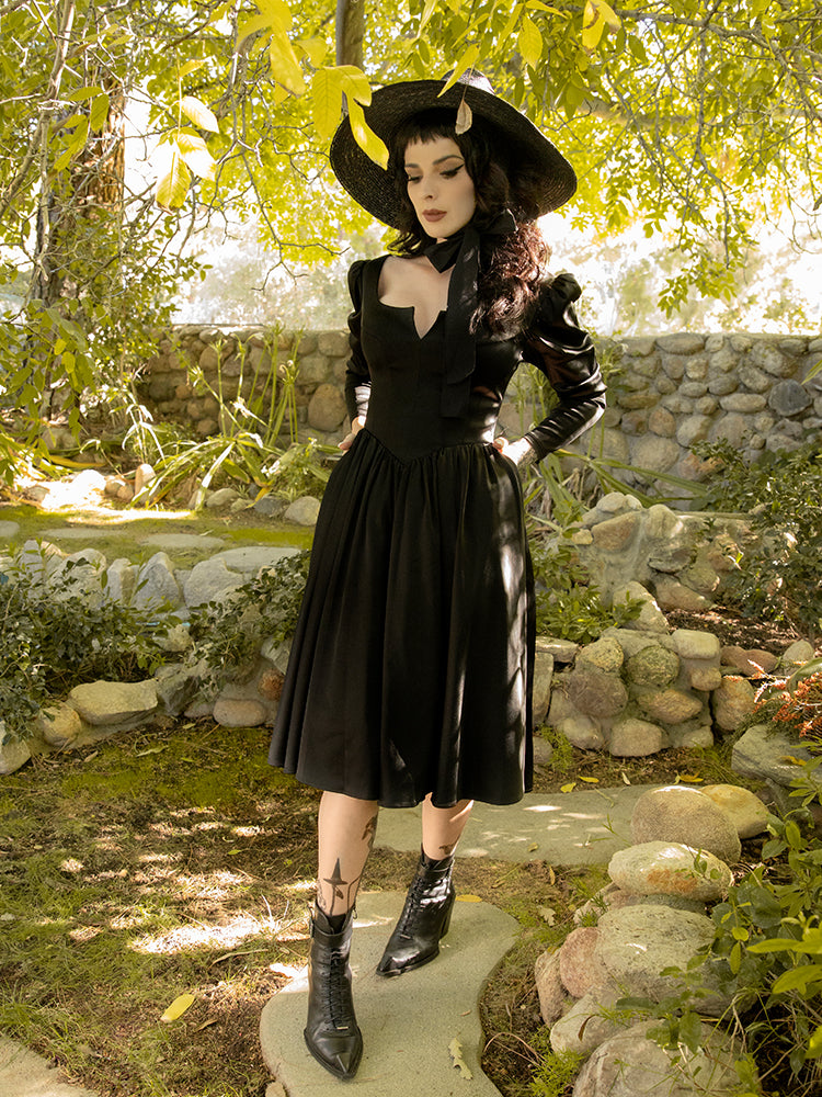 Walking on a stone pathway amongst a garden setting, Stephanie models the Cottage Witch Dress in Japanese Black Satin from goth dress maker La Femme en Noir.