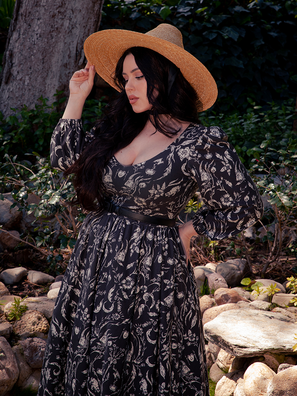 Within the depths of the mystic forest, a breathtaking dark-haired beauty emerges, veiled in the mesmerizing Dark Forest Dress, donning the haunting Cottage Witch Toile Print, a masterpiece by the esteemed goth dress maker La Femme en Noir.