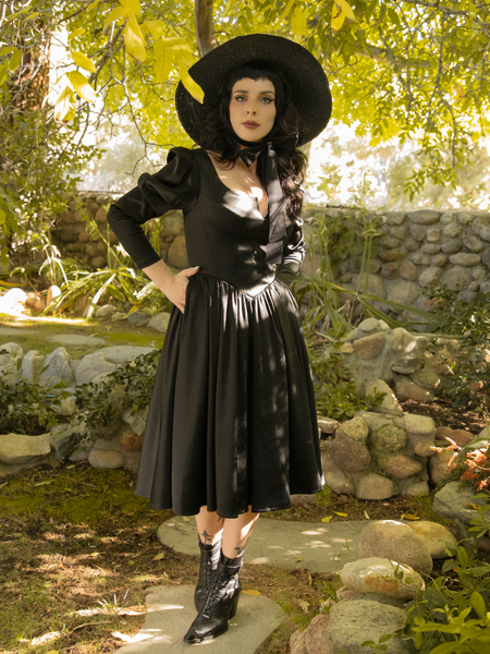 Looking into the camera, Stephanie places her hands in the pockets of her Cottage Witch Dress in Japanese Black Satin from gothic clothing company La Femme en Noir.