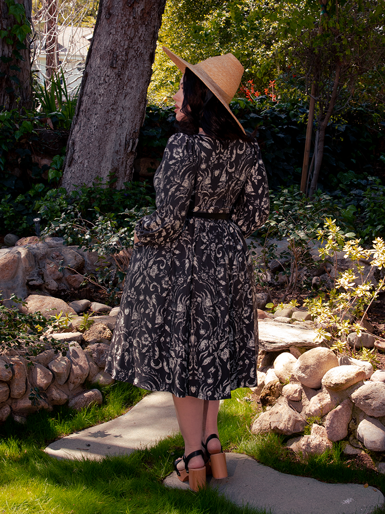 Within the mysterious forest sanctuary, a captivating brunette beauty enchants the realm, draped in the enchanting Dark Forest Dress embellished with the ethereal Cottage Witch Toile Print, an alluring ensemble from goth dress maker La Femme en Noir.