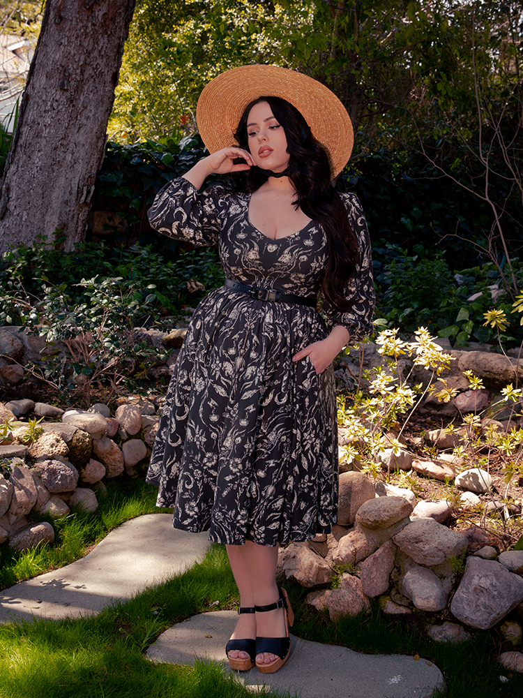 Amongst the eerie foliage of the forest, a striking brunette model captivates the imagination, draped in the alluring Dark Forest Dress adorned in the hauntingly beautiful Cottage Witch Toile Print, a creation by La Femme en Noir, the gothic fashion virtuoso.