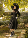 Striking a pose with one hand near her face and the other tucked into a pocket on her Cottage Witch Dress in Japanese Black Satin, Stephanie models the latest goth clothing release from La Femme en Noir.
