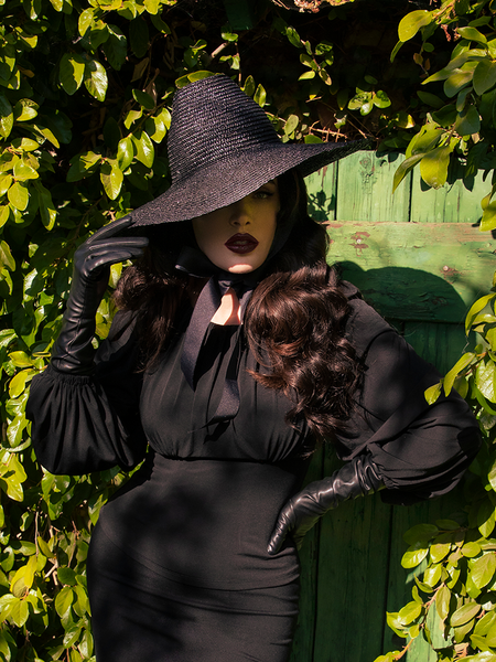 Micheline Pitt in an all black outfit featuring the Cottage Witch Hat in Black along with an all black gothic long sleeve dress and vinyl gloves.