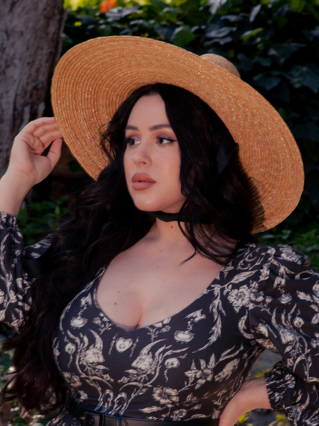 The Cottage Witch Hat in Natural from La Femme en Noir adds an enchanting touch to the dark haired female model's gothic clothing outfit, making her look stunning.
