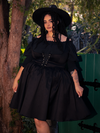 Lost in the mysterious ambiance of the forest, a stunning fair-skinned brunette figure embodies gothic glamour, showcasing the captivating Cottage Corset Pinafore in Black, an exquisite addition to your gothic dress repertoire.