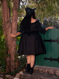 Within the shady sanctuary of the forest, a captivating fair-skinned brunette model personifies gothic style, wearing the enchanting Cottage Corset Pinafore in Black, a striking accent piece that complements your gothic dress collection.