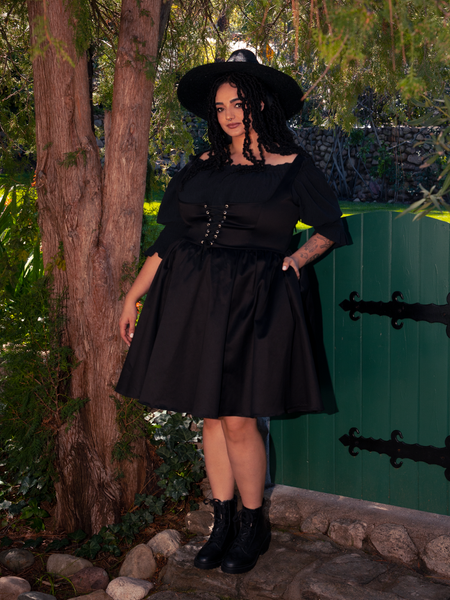 Amidst the enchanting forest backdrop, a stunning fair-skinned brunette muse embraces gothic fashion with poise, draped in the elegant Cottage Corset Pinafore in Black, a versatile addition to your gothic dress wardrobe.