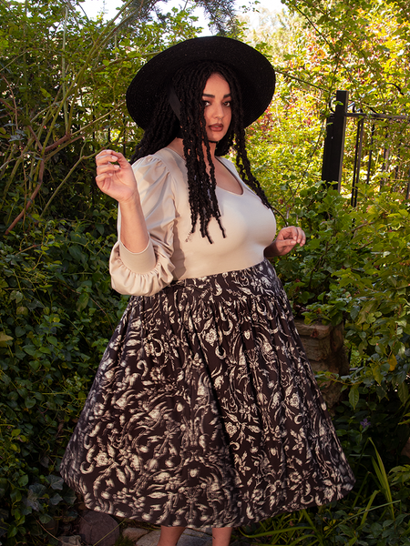 The stunning female model confidently displays the Gothic Tales Skirt in Cottage Witch Toile Print from the gothic clothing brand La Femme en Noir.