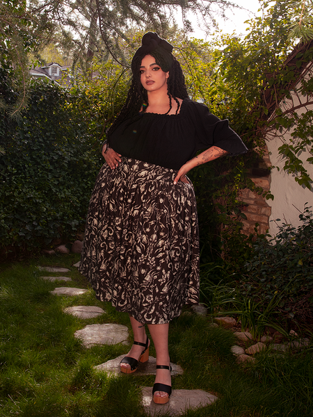 With poise and charm, the beautiful female model displays the Gothic Tales Skirt in Cottage Witch Toile Print from the gothic clothing brand La Femme en Noir.