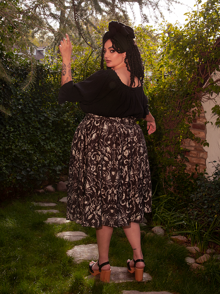 Standing out with elegance, the female model shows off the Gothic Tales Skirt in Cottage Witch Toile Print from the gothic clothing brand La Femme en Noir.