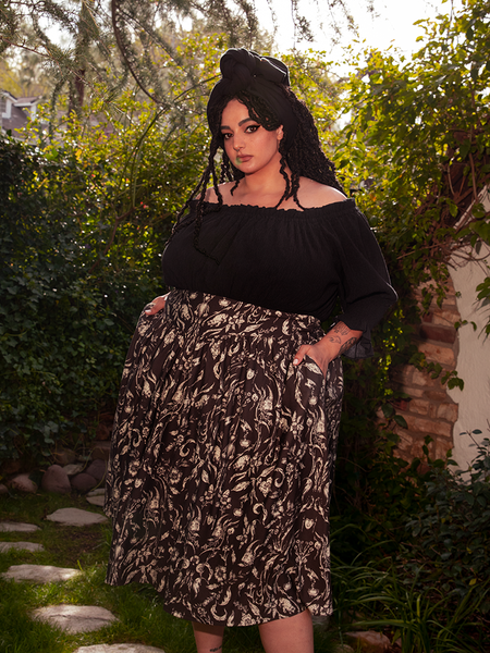 Posing with confidence, the stunning female model presents the Gothic Tales Skirt in Cottage Witch Toile Print from the gothic clothing brand La Femme en Noir.