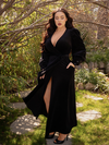 Rachel Sedory standing in a garden looking down while wearing the Crawford Hostess Wrap Gown in Black.