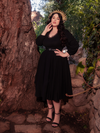 Embracing the serene ambiance of a forest setting, a striking brunette model exudes allure as she adorns herself with the bewitching Dark Forest Dress in Black, a remarkable creation by the gothic fashion brand, La Femme en Noir.