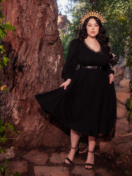 With a backdrop of a forest's natural splendor, a stunning brunette model stands poised, clad in the alluring Dark Forest Dress in Black from the gothic fashion label, La Femme en Noir.