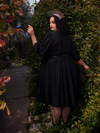 Standing amidst a picturesque forest, a radiant brunette model showcases her beauty, adorned in the Dark Forest Dress in Black from La Femme en Noir, a renowned gothic fashion brand.