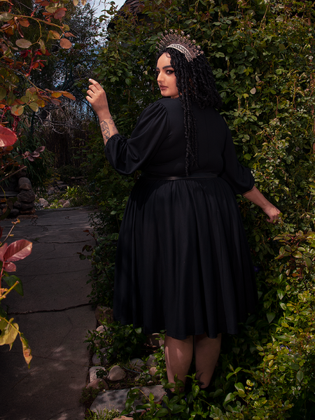 Standing amidst a picturesque forest, a radiant brunette model showcases her beauty, adorned in the Dark Forest Dress in Black from La Femme en Noir, a renowned gothic fashion brand.