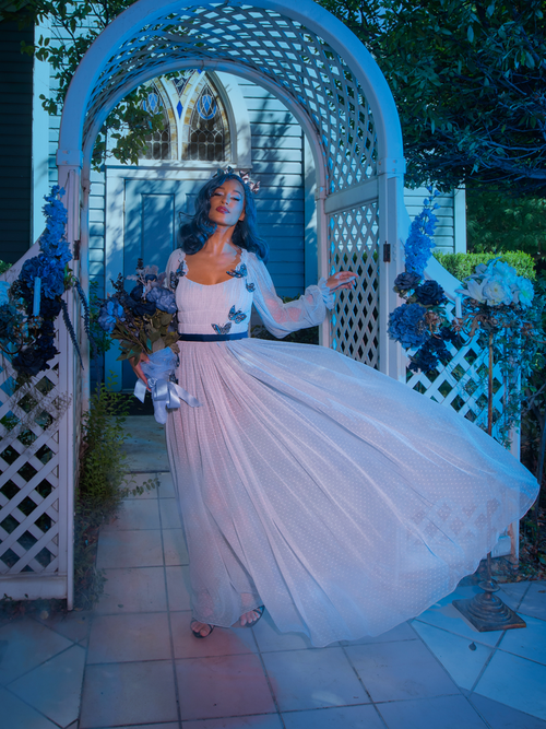Full length image of model standing in a dimly lit garden wearing the Tim Burton's CORPSE BRIDE™ Emily Butterfly Gown in Celestial Blue. 