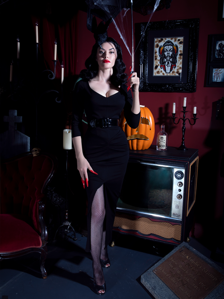 Heather standing a retro spooky living room wearing the Glamour Ghoul Gown from gothic clothing company La Femme en Noir.