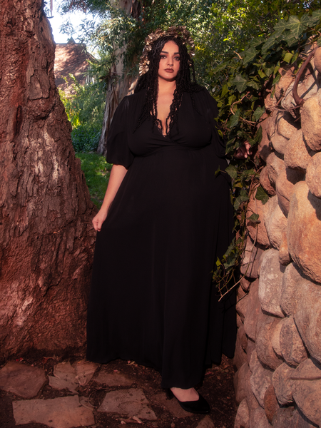 Model standing next to an old tree and a rock wall while modeling the Odyssey Maxi Dress in Black from gothic clothing brand La Femme en Noir.