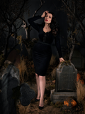 Full length image of Micheline Pitt leaning on a tombstone while wearing a black gothic style dress. 