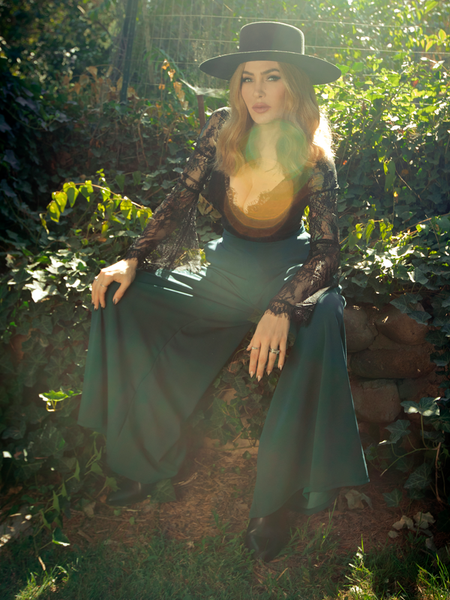 Micheline Pitt sits in a lush garden while modeling the Black Widow palazzo pants in hunter green.