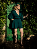 A full length shot of Micheline Pitt, with her hand in her pocket standing in a garden, models the Black Widow tap shorts in hunter green from La Femme En Noir.