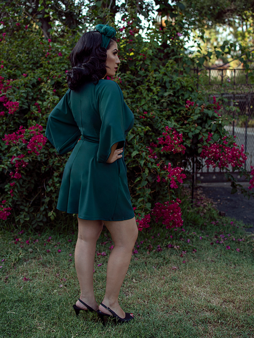 A back shot of Erika, with her hands on her hips in a garden, models the Black Widow tap shorts in hunter green from La Femme En Noir.