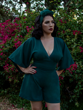 Erika, with her hands on her hips in a garden, models the Black Widow tap shorts in hunter green from La Femme En Noir.