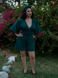A full length shot of Erika, with her hands on her hips in a garden, models the Black Widow tap shorts in hunter green from La Femme En Noir.