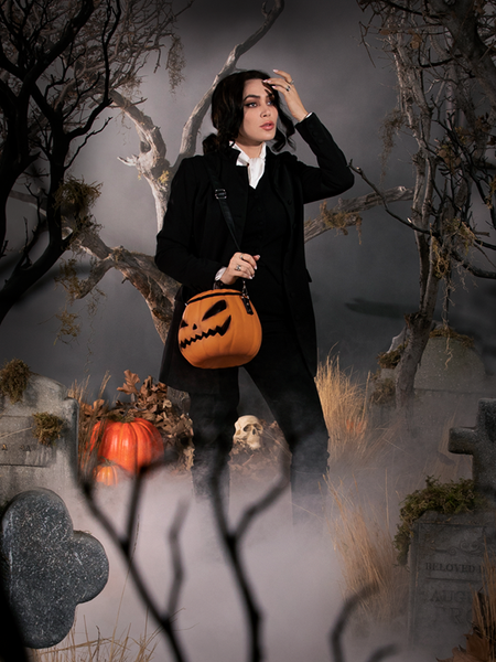 Reaching up to move the hair away from her brow, Micheline Pitt trudges through a fog covered graveyard scene while carrying the Sleepy Hollow Pumpkin Bag. 