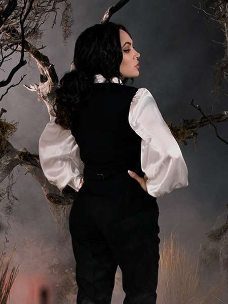 Micheline Pitt, turned away from the camera, looks back slightly over her shoulder while modeling the Sleepy Hollow Ichabod Vest in Black over a flowy white shirt and black pants. 