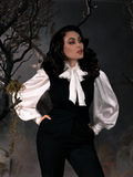 With her hands tucked into her pockets, Micheline Pitt gazes off while standing in a spooky graveyard setting while wearing the Victorian Blouse in Ivory.