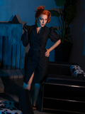 Full length shot of red-haired female model standing in a spooky living room wearing the BEETLEJUICE™ Delia Dress.