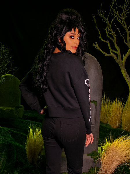 Back shot of a female model in an all black goth style outfit highlighted by the BEETLEJUICE™ Lydia Strange Long Sleeve Tee.