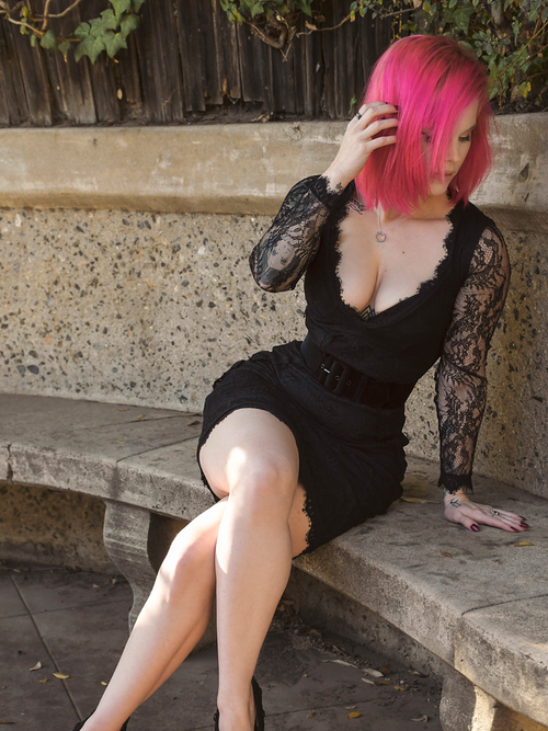 Mackenzie wearing the La Dentelle Dress in Black while sitting on a bench in front of a green, wooded area.